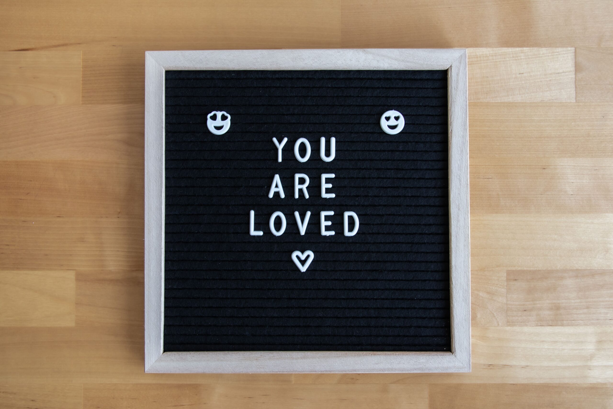 Black letter board on a wooden table with white letters that read “You are loved.”
